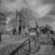 A black and white photograph of St. Giles' Church in Hartington, Derbyshire, capturing the historic atmosphere of the churchyard with aged gravestones and a dramatic sky.