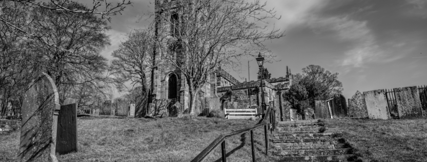 A black and white photograph of St. Giles' Church in Hartington, Derbyshire, capturing the historic atmosphere of the churchyard with aged gravestones and a dramatic sky.
