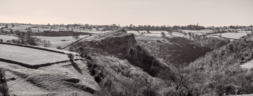 A monochrome landscape of the Peak District, featuring the dramatic limestone cliffs near Thor’s Cave, surrounded by rolling hills and dry stone walls.