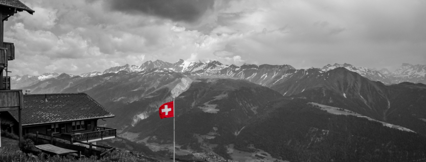 Black and white alpine landscape with a Swiss flag in red standing above a valley and distant mountain range under dramatic clouds