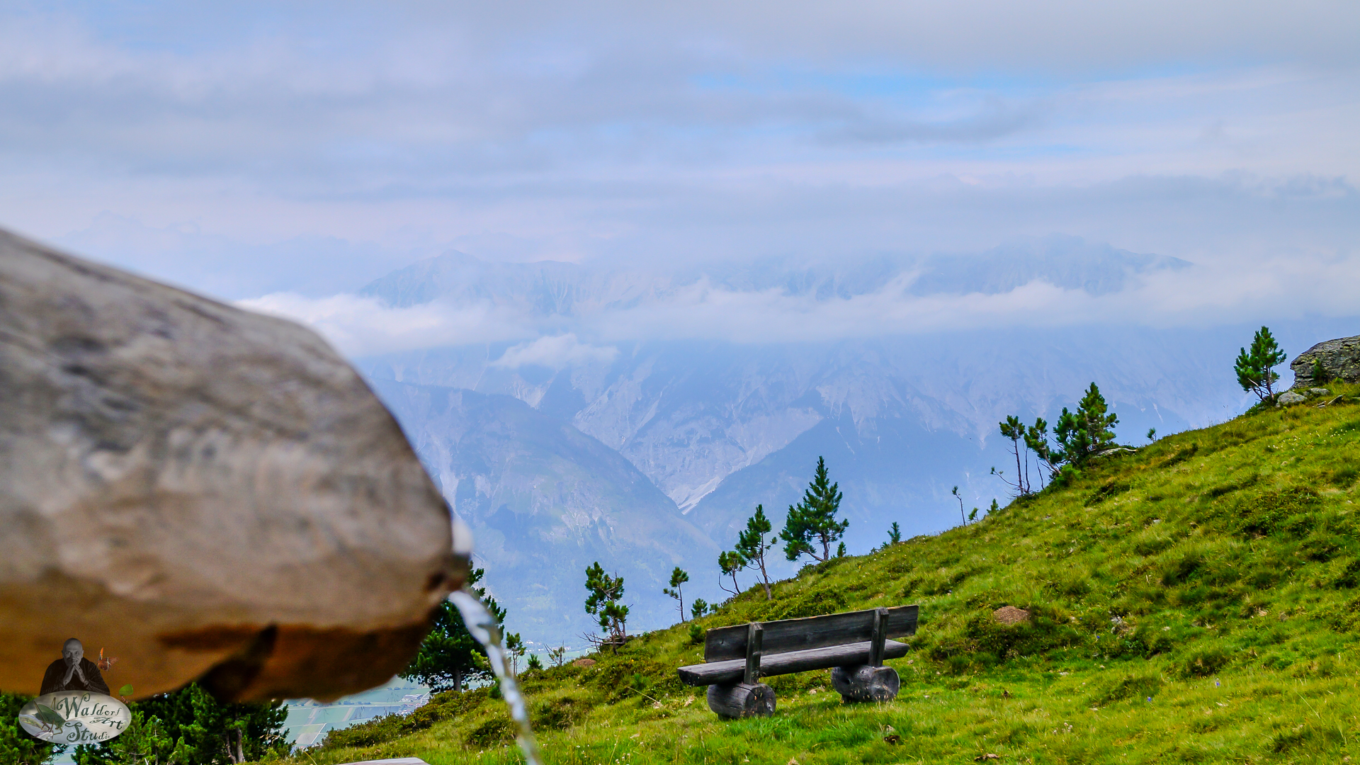 A wooden bench on a grassy alpine slope faces distant misty mountains, while water flows gently from a stone fountain in the foreground.